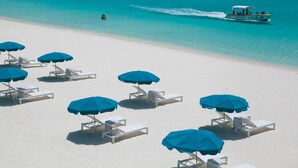 Una playa con sillones blancos y sombrillas azules, un barco en el agua y un mar turquesa cristalino.