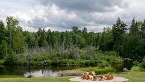 Un entorno junto al lago con una zona de estar circular, con sillas y una mesa de madera, rodeada de césped y un bosque frondoso.