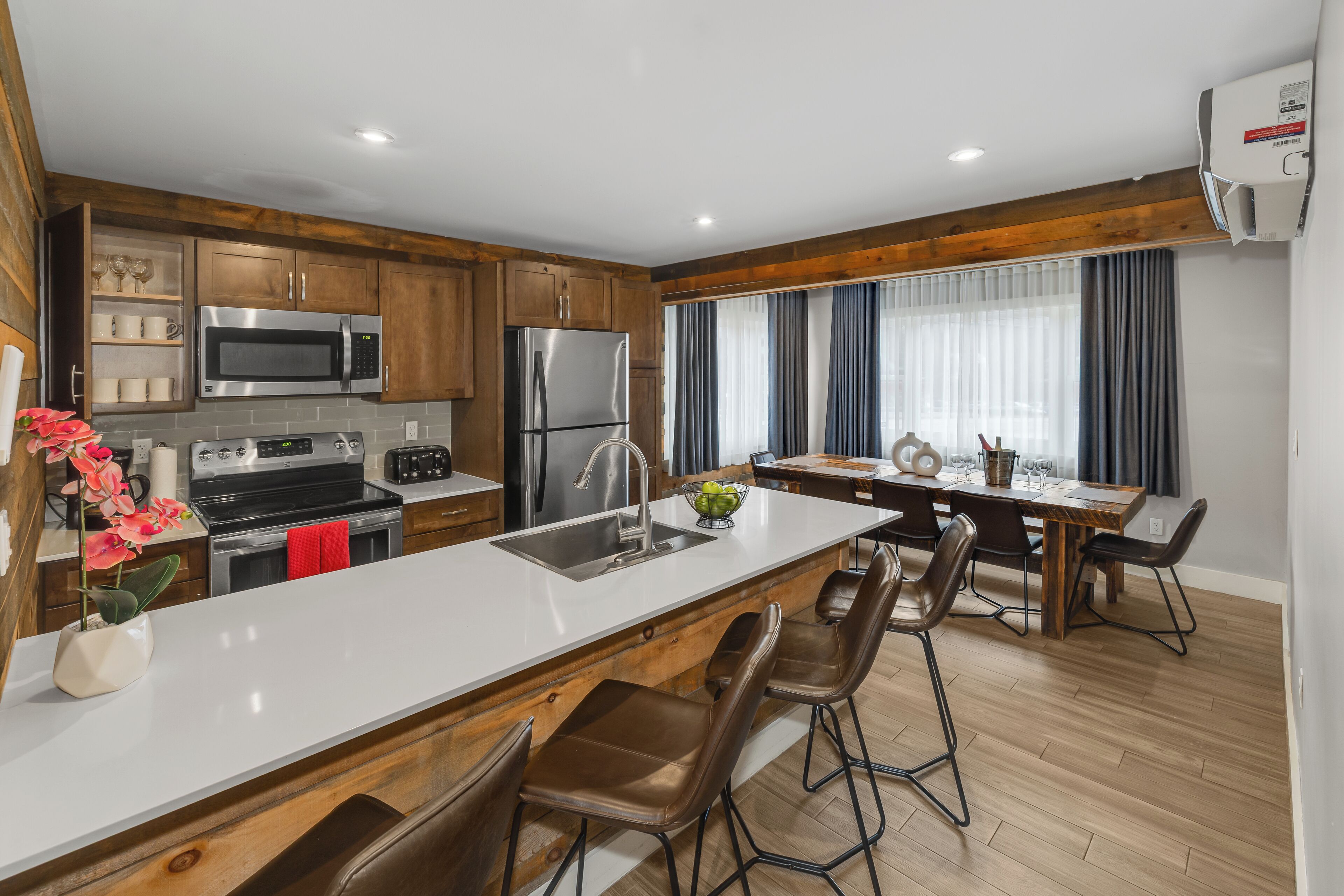 A modern kitchen with wooden cabinets, stainless steel appliances, a central island with bar stools, and a dining area with a table and chairs.