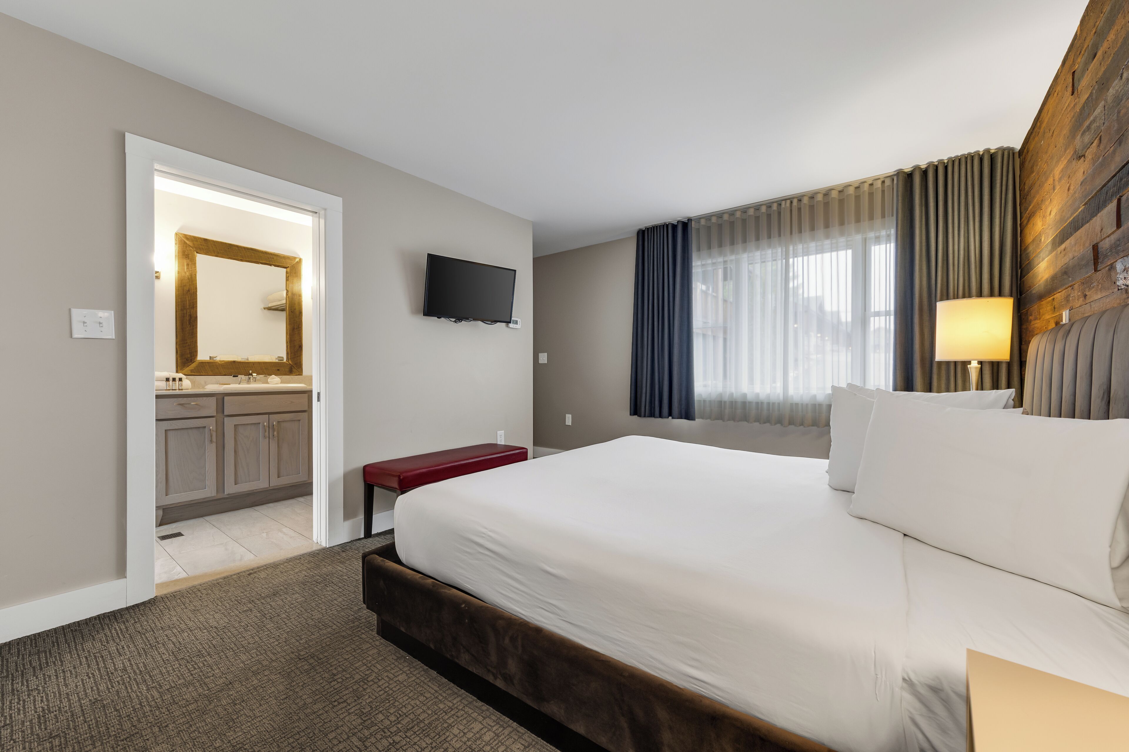 Ein Hotelzimmer mit einem großen Bett, einem an der Wand befestigten Fernseher, einem Holzkopfteil und einem Fenster mit Vorhängen.
