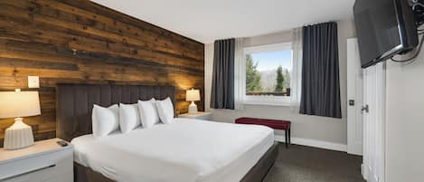 Habitación de hotel con cabecera de madera, una cama grande, televisor de pantalla plana en la pared, ventana con cortinas y una pequeña mesa con un banco rojo.