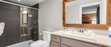 A modern bathroom with a glass-enclosed shower, a white toilet, a large mirror with a wooden frame, and a vanity with a sink and cabinets.