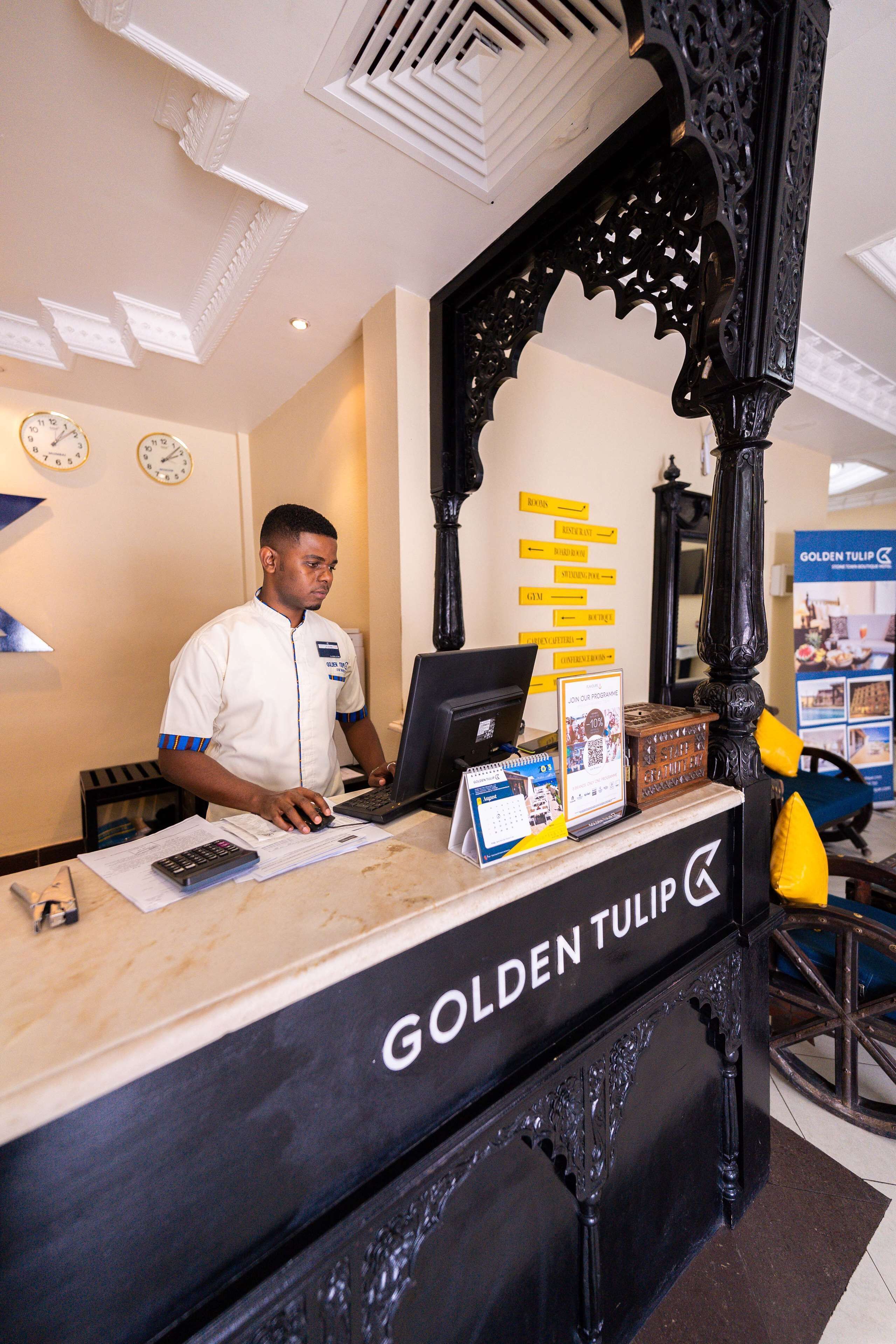 A hotel reception desk with a man working on a laptop, a clock, and a sign for "Golden Tulip."