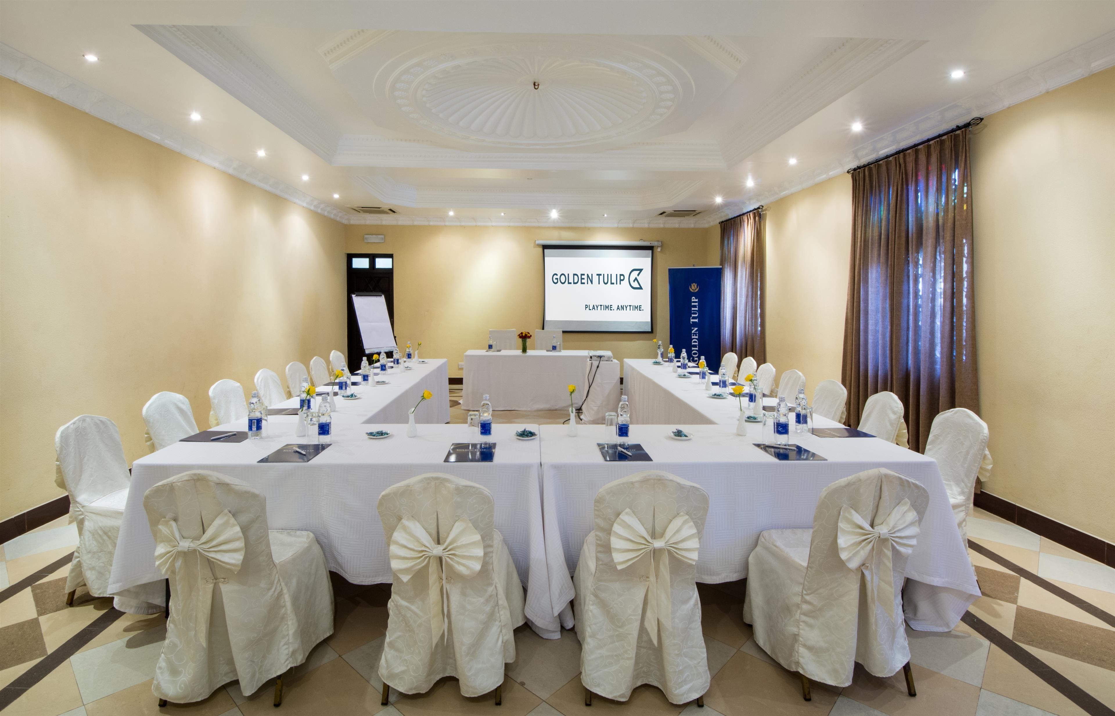 A conference room with a long table, white chairs with bows, a projector screen, and a banner reading "GOLDEN TULIP PLAYTIME, ANYTIME."