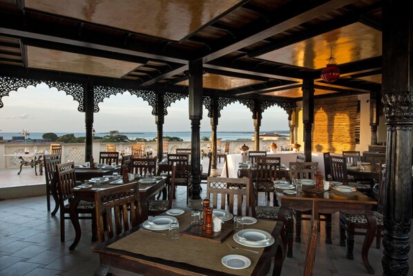An outdoor dining area with wooden tables and chairs, a view of the sea, and ornate architectural details.
