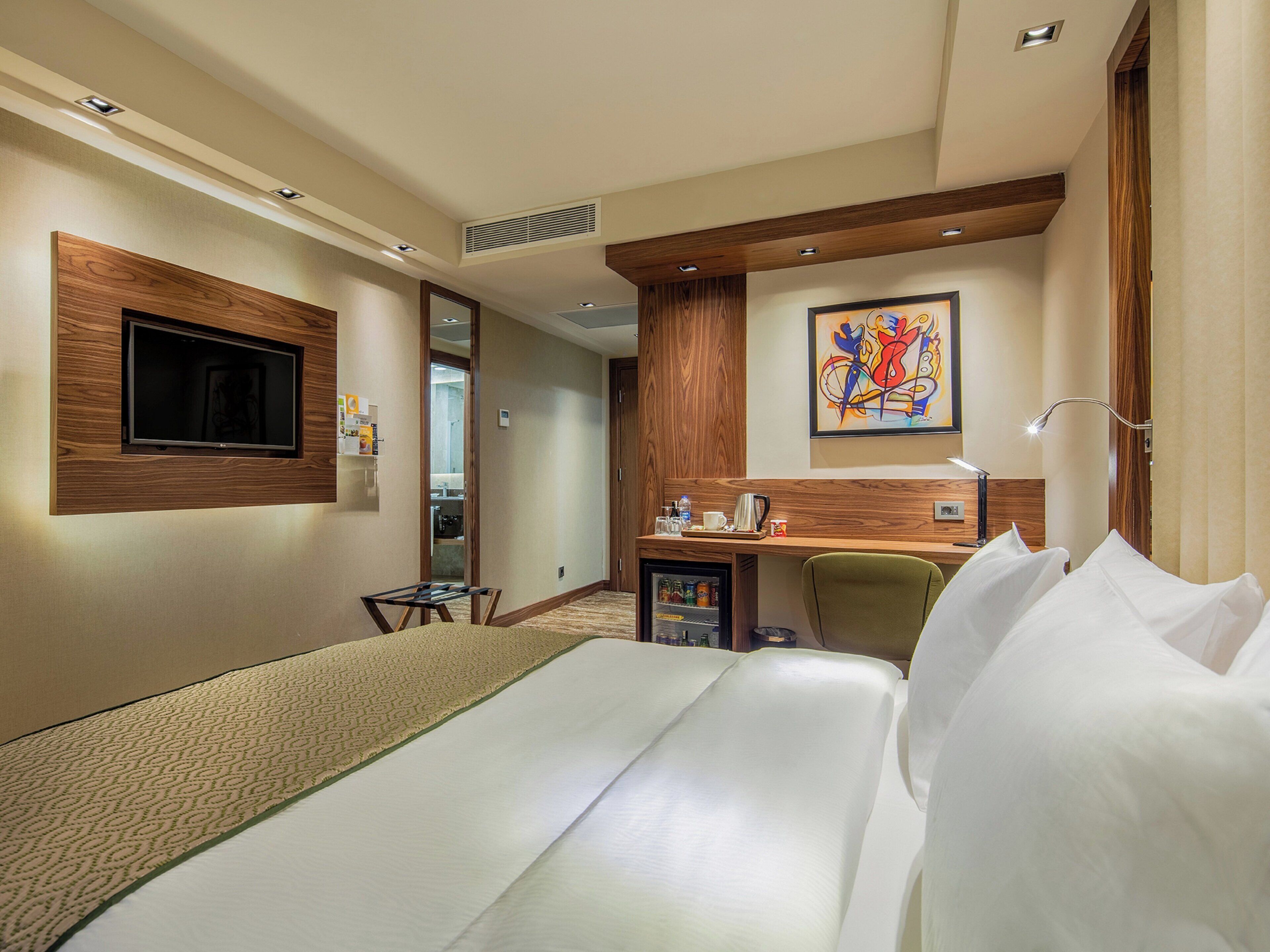 A hotel room with a bed, a desk, a chair, a television, a painting, and a minibar.