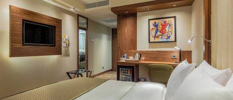 A hotel room with a bed, a desk, a chair, a television, a painting, and a minibar.