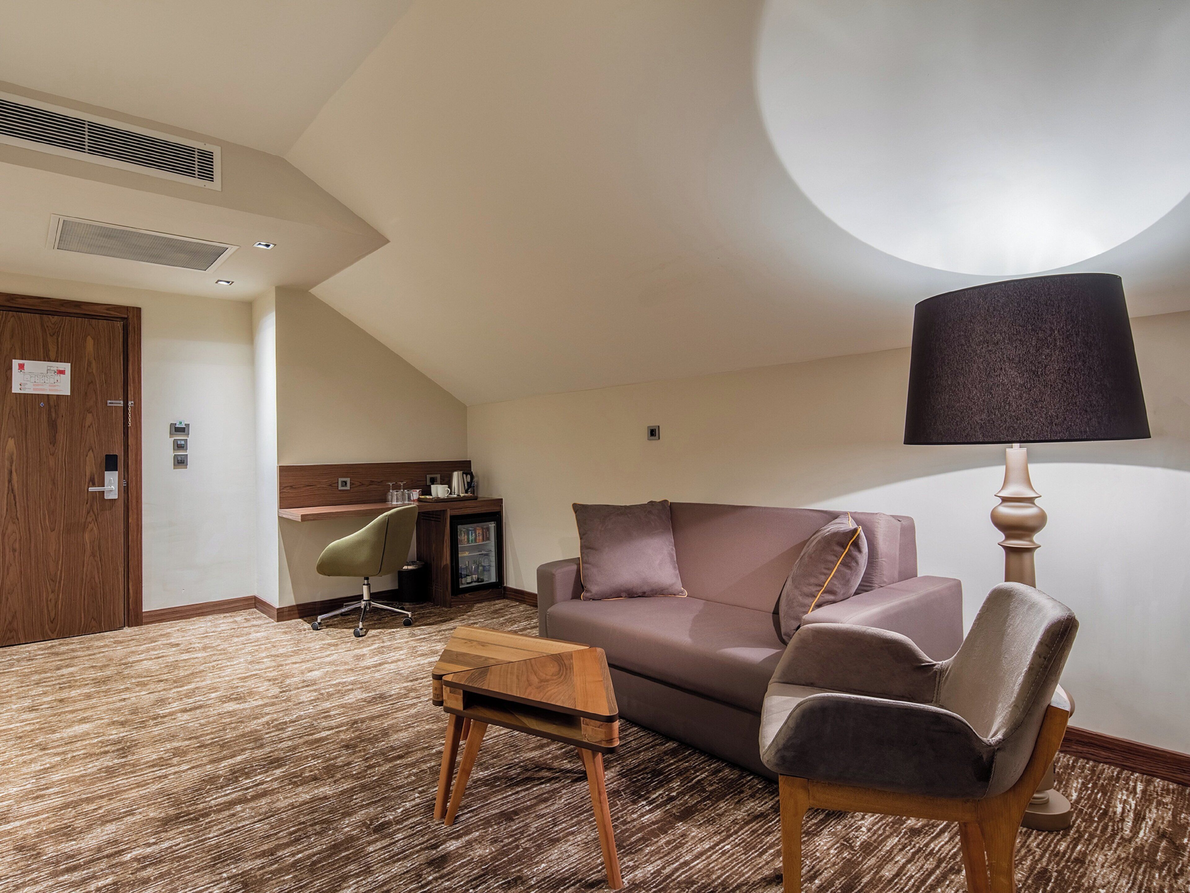 Ein Hotelzimmer mit einer Couch, einem kleinen Tisch, einer Lampe, einem Sessel, einem Schreibtisch und einem Kühlschrank.
