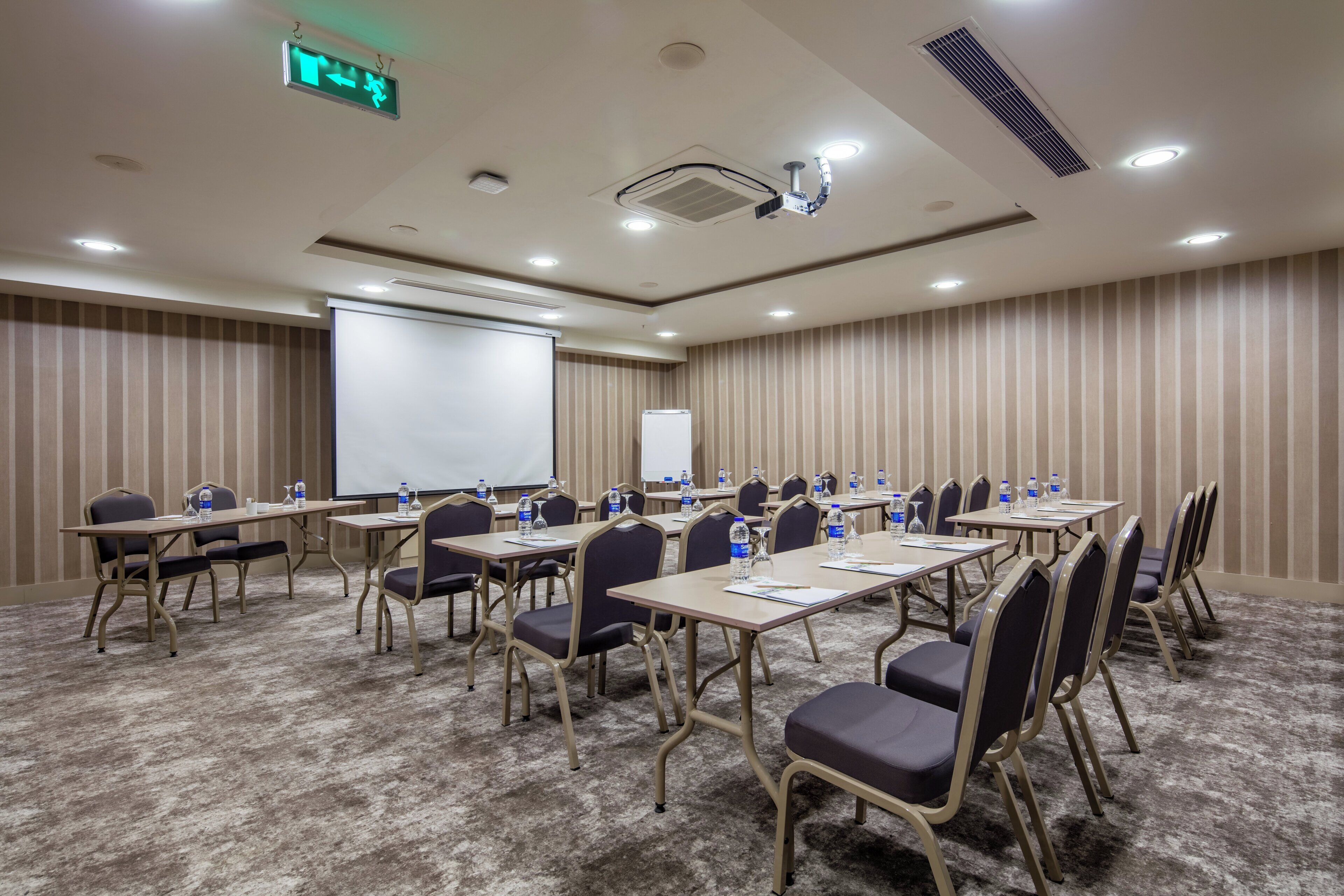 Une salle de conférence équipée de plusieurs tables, de chaises, d’un écran de projection et d’un tableau blanc.