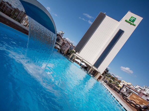 A hotel with a large swimming pool, a modern building with a green logo, and a clear blue sky.