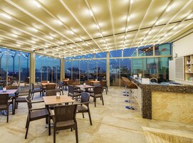 A spacious indoor area with tables and chairs, large windows, and a ceiling with lights.