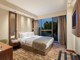 A hotel room with a large bed, a desk, a chair, a mirror, a TV, and a window with curtains.
