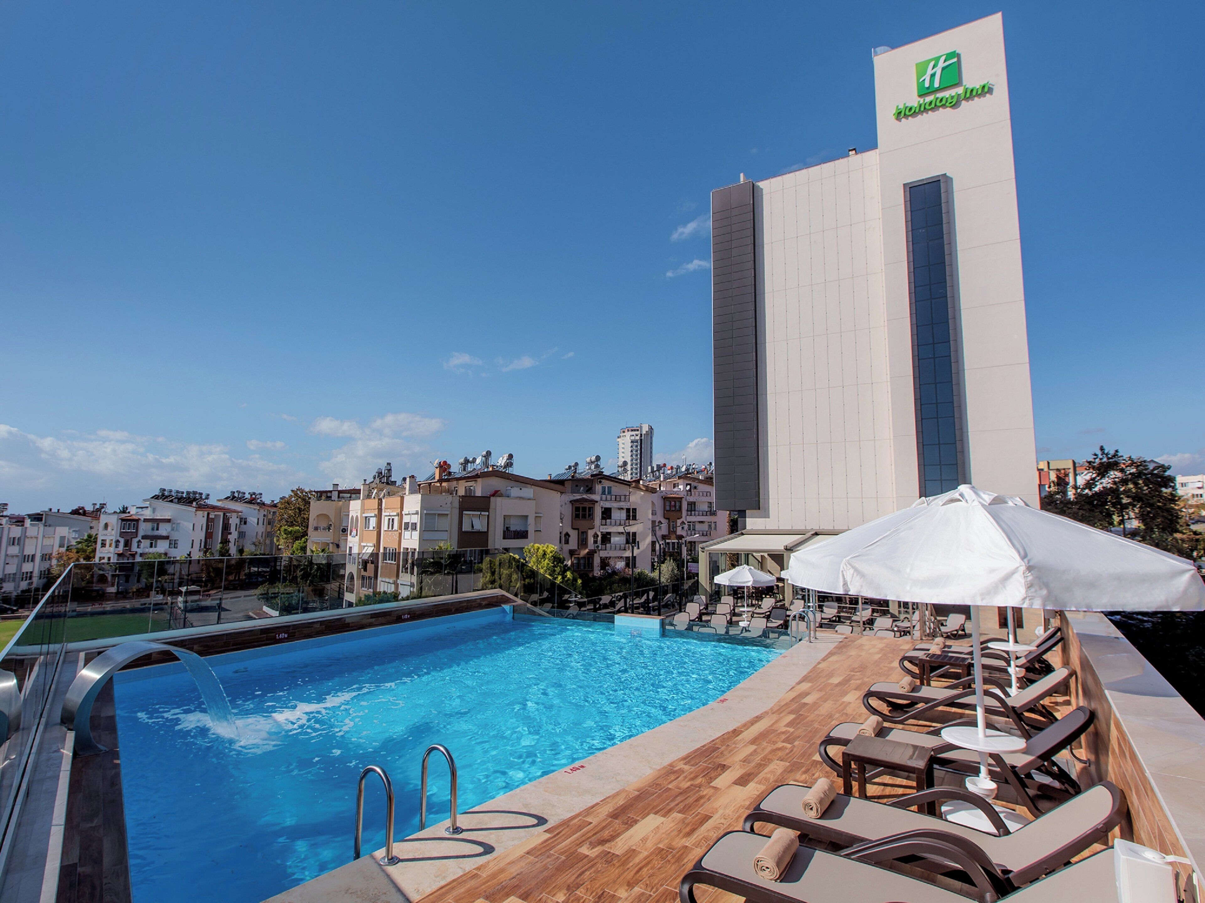 Piscina en la azotea con sillones de descanso y sombrillas, con vistas a un paisaje urbano donde destaca un edificio con logotipo verde.
