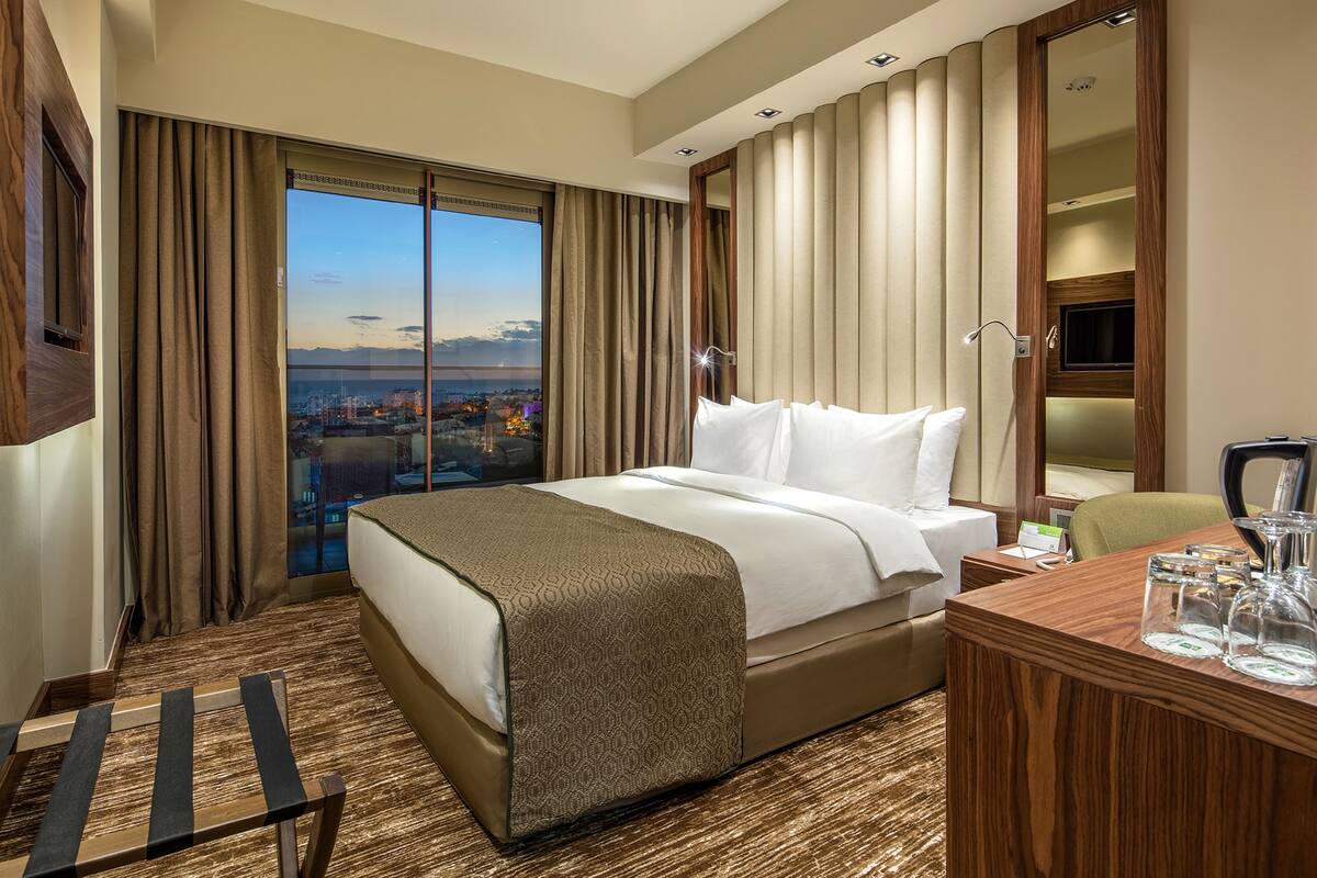 A hotel room with a large bed, a desk with a chair, a mirror, and a view of a cityscape through large windows.