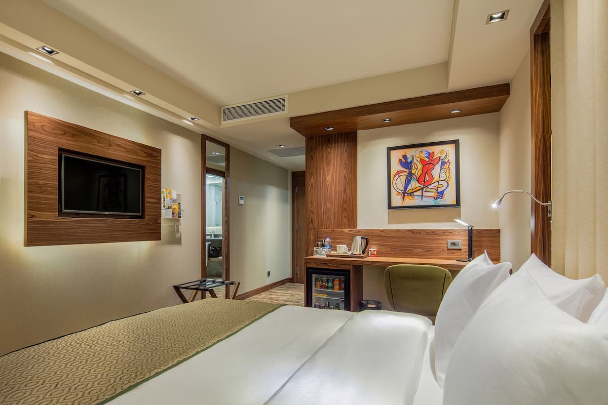 A hotel room with a bed, a desk, a chair, a TV, a painting, and a minibar.