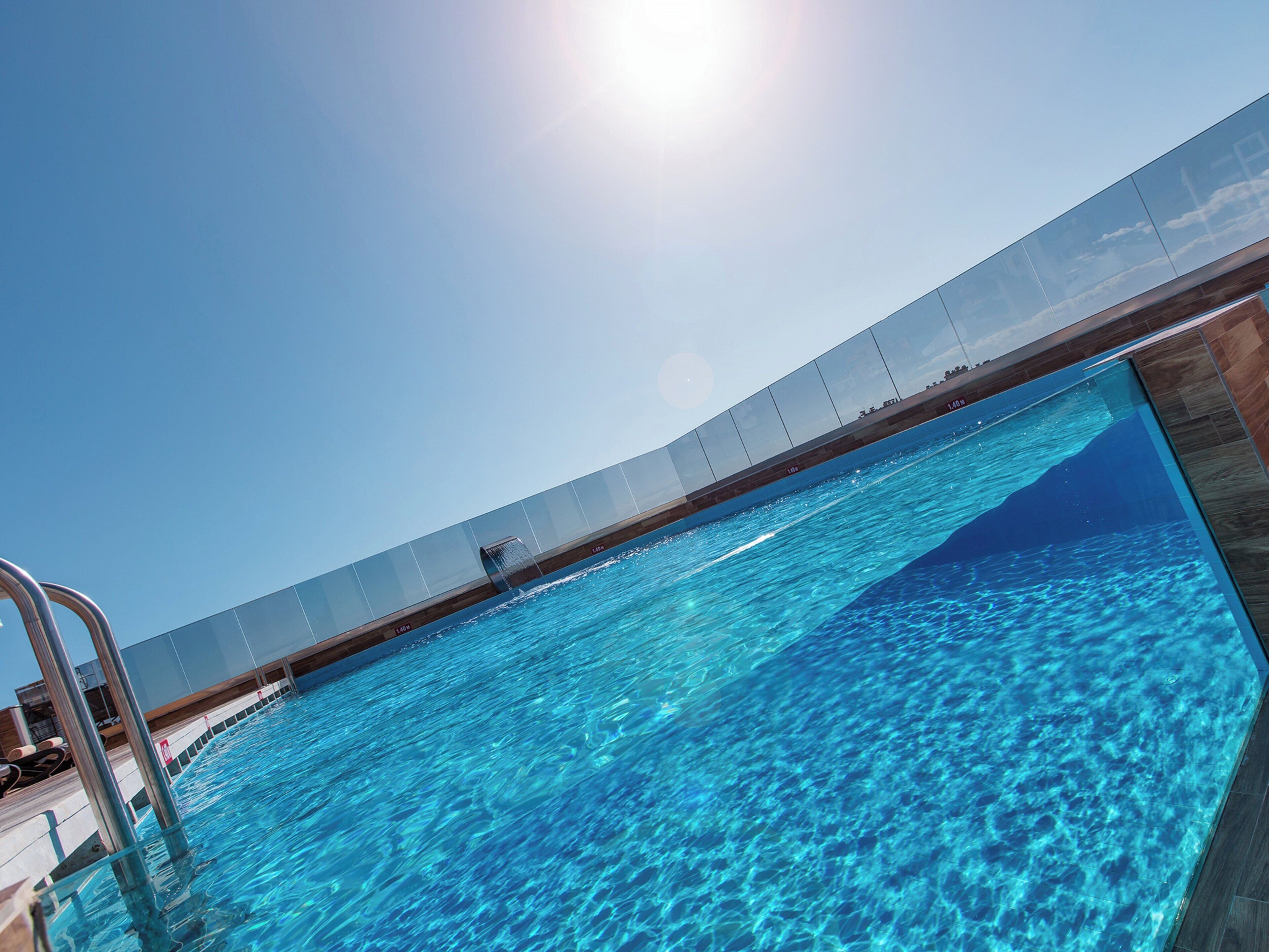 A clear blue pool with a glass ceiling, a metal ladder, and a wooden deck.