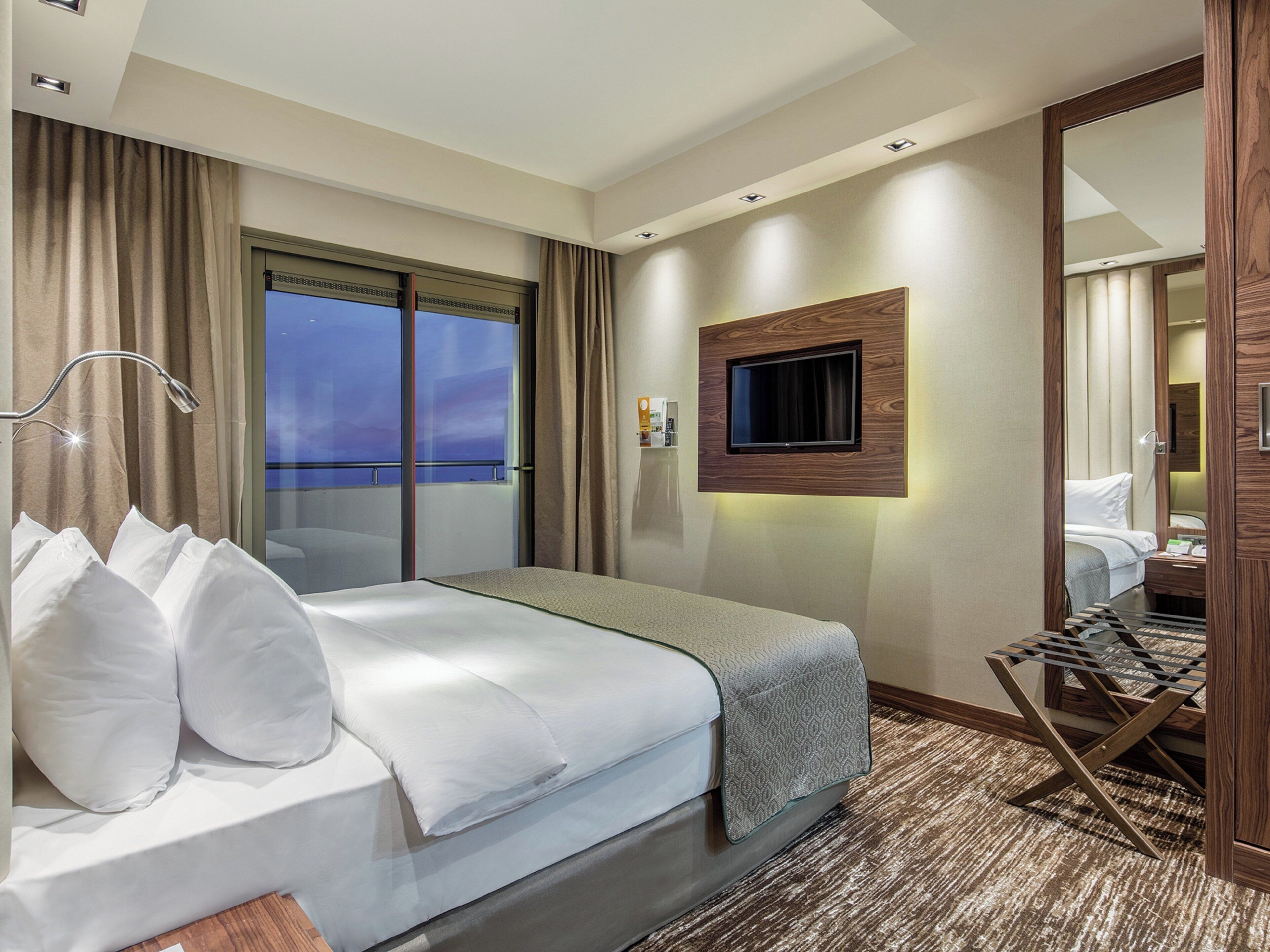Een hotelkamer met een bed, een aan de muur bevestigde televisie, een raam met gordijnen en uitzicht op de omgeving.
