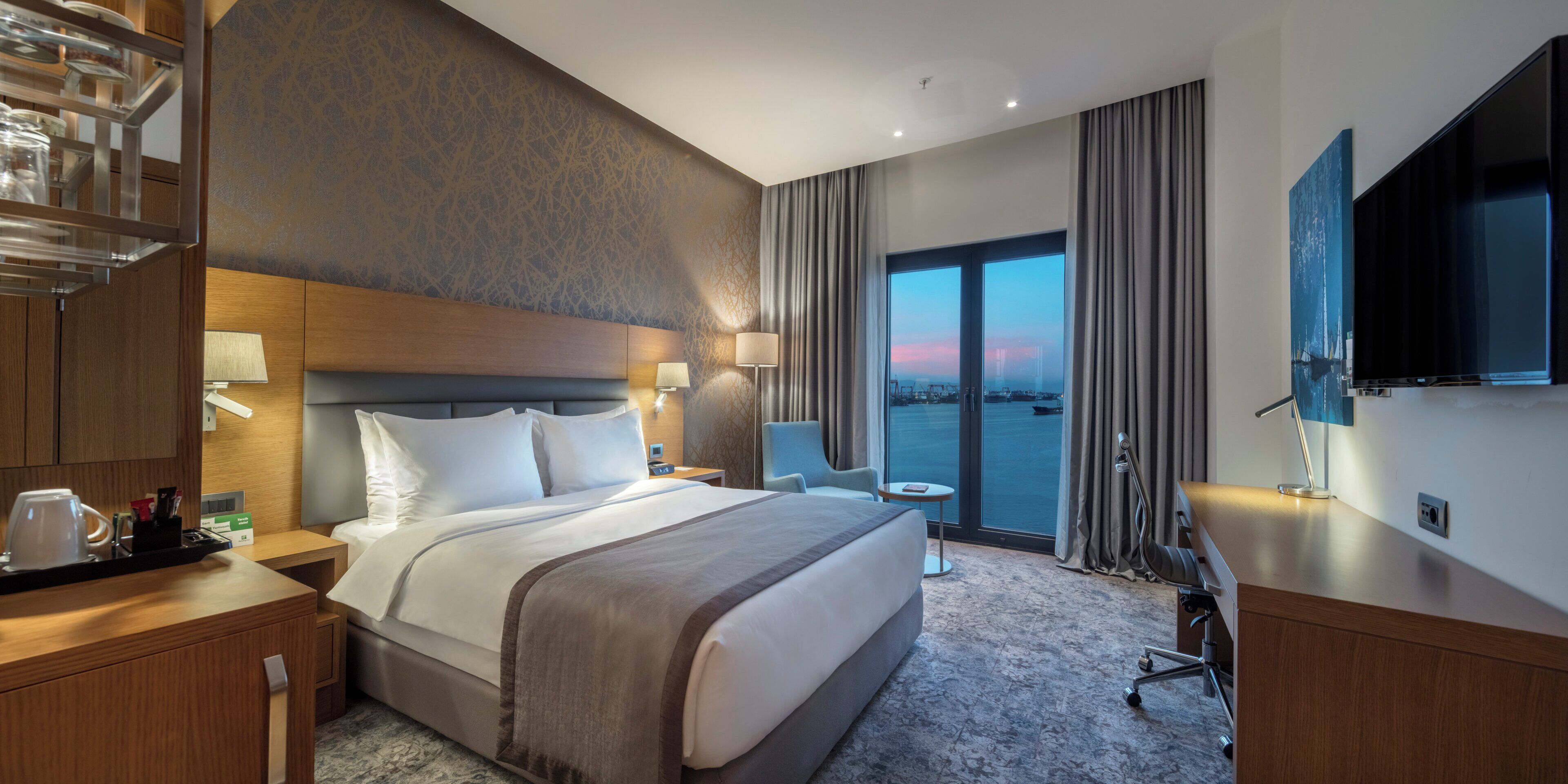 A hotel room with a large bed, a desk with a lamp, a chair, a TV, and a view of the water.
