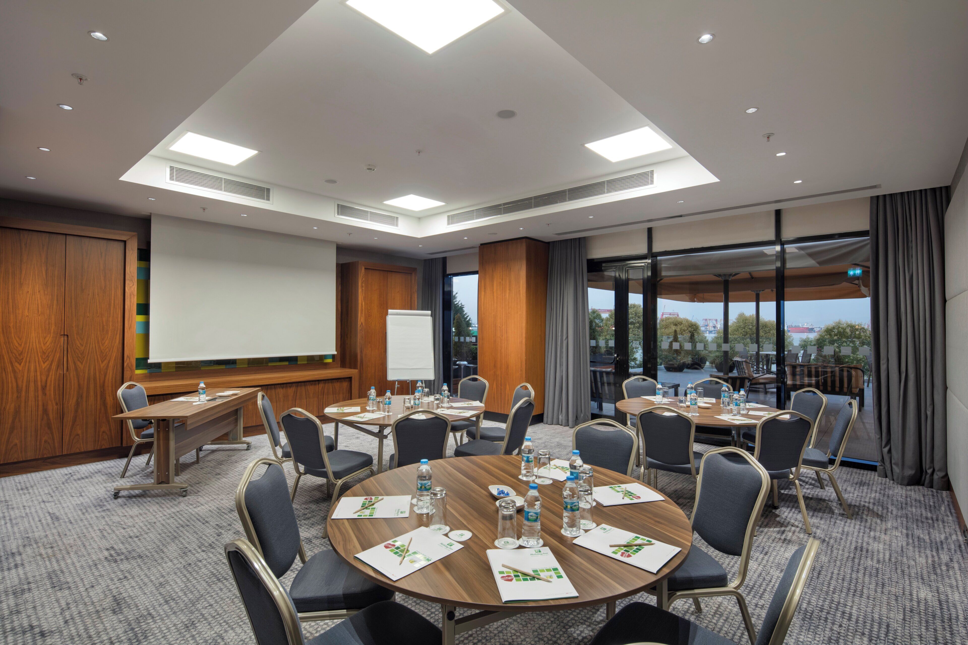 A conference room with a large table, chairs, a projector screen, and a view of the outdoors.