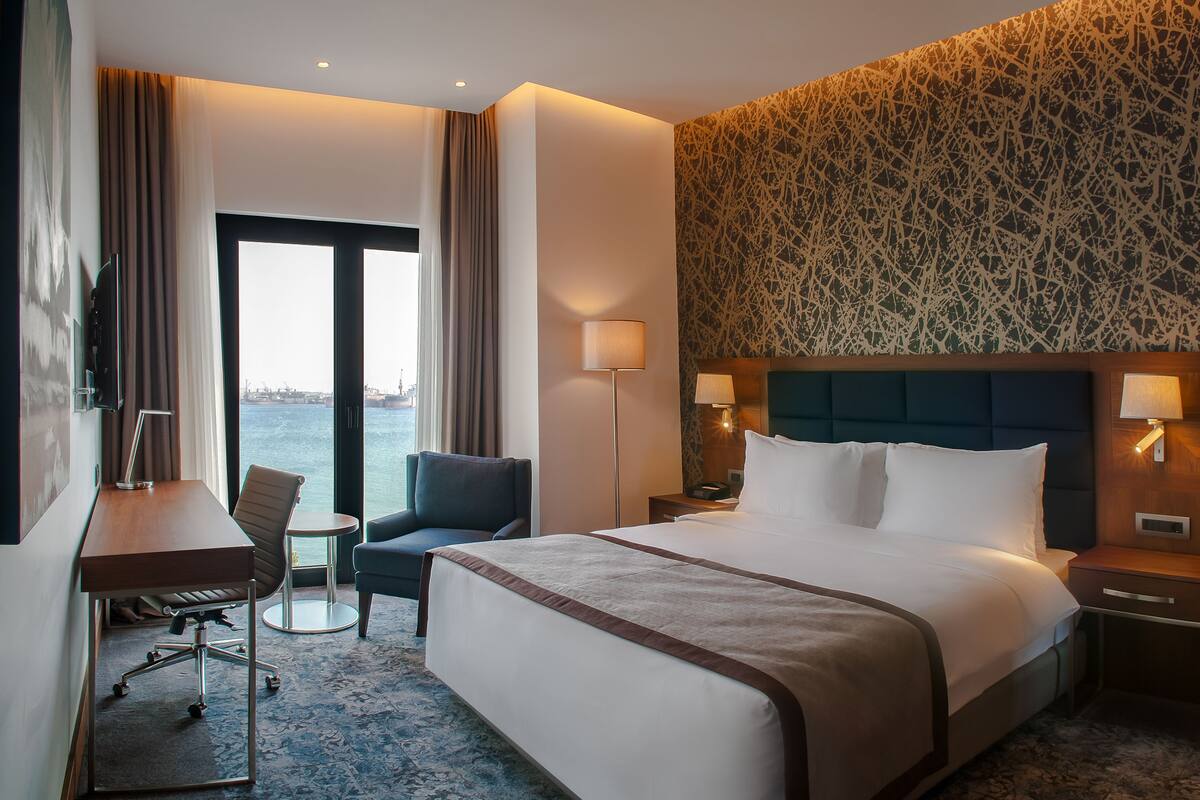 A hotel room with a large bed, a desk, a chair, a small table, and a view of the water.