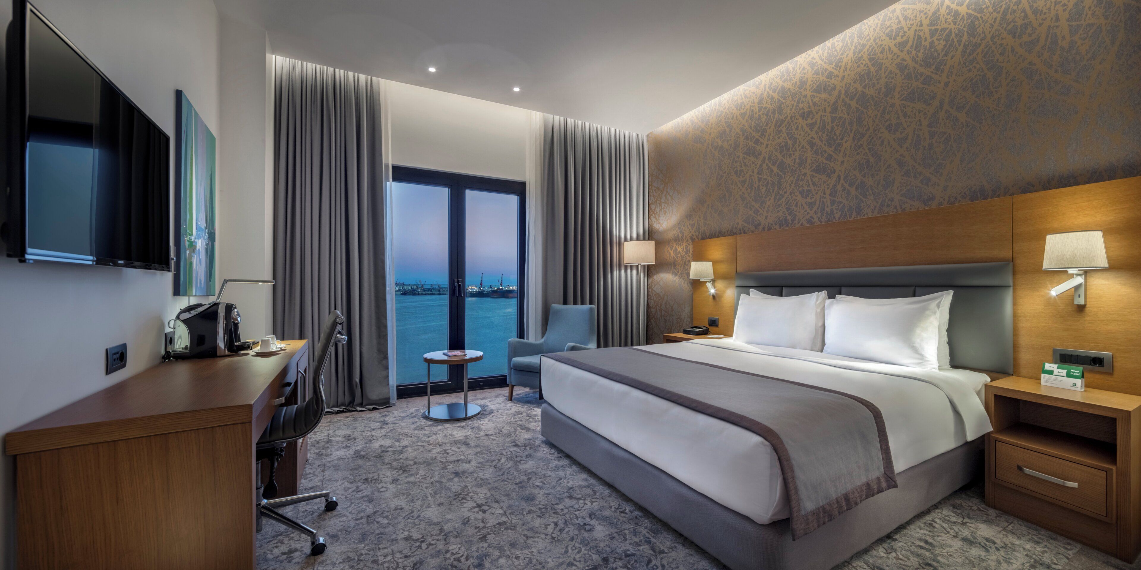 Hotelkamer met een groot bed, een bureau met stoel, een televisie, uitzicht op het water en een kamerwand met een patroon.
