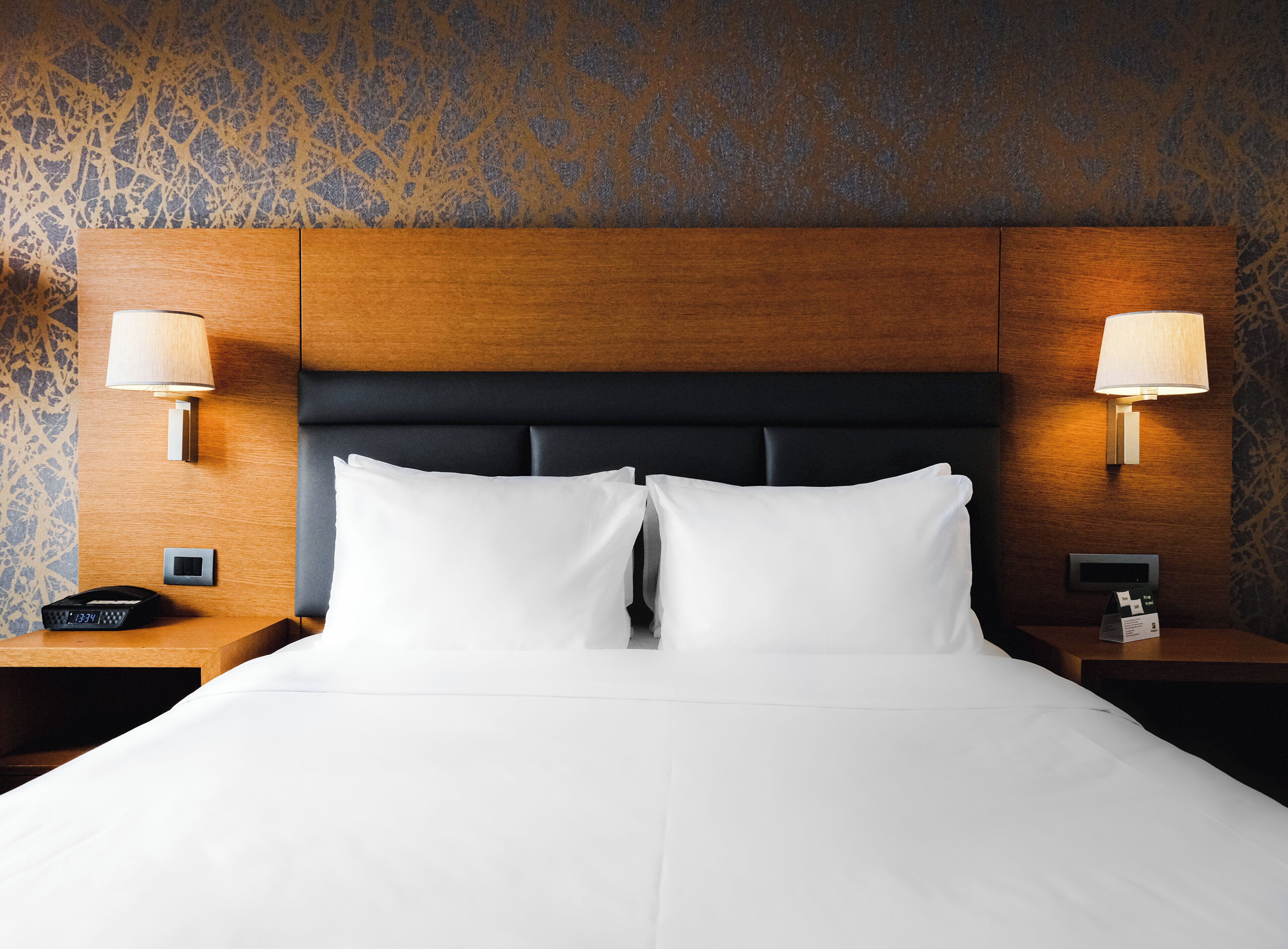 A hotel room with a bed, two bedside lamps, a nightstand with a phone, and a wall clock.