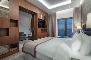 A modern hotel room with a large bed, a seating area with two armchairs, a television, and a view of the cityscape through the window.