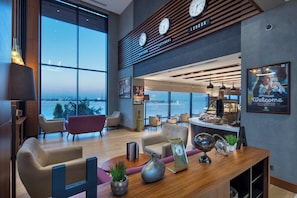 A modern hotel lobby with a large window offering a view of the cityscape, a reception desk, and seating area.