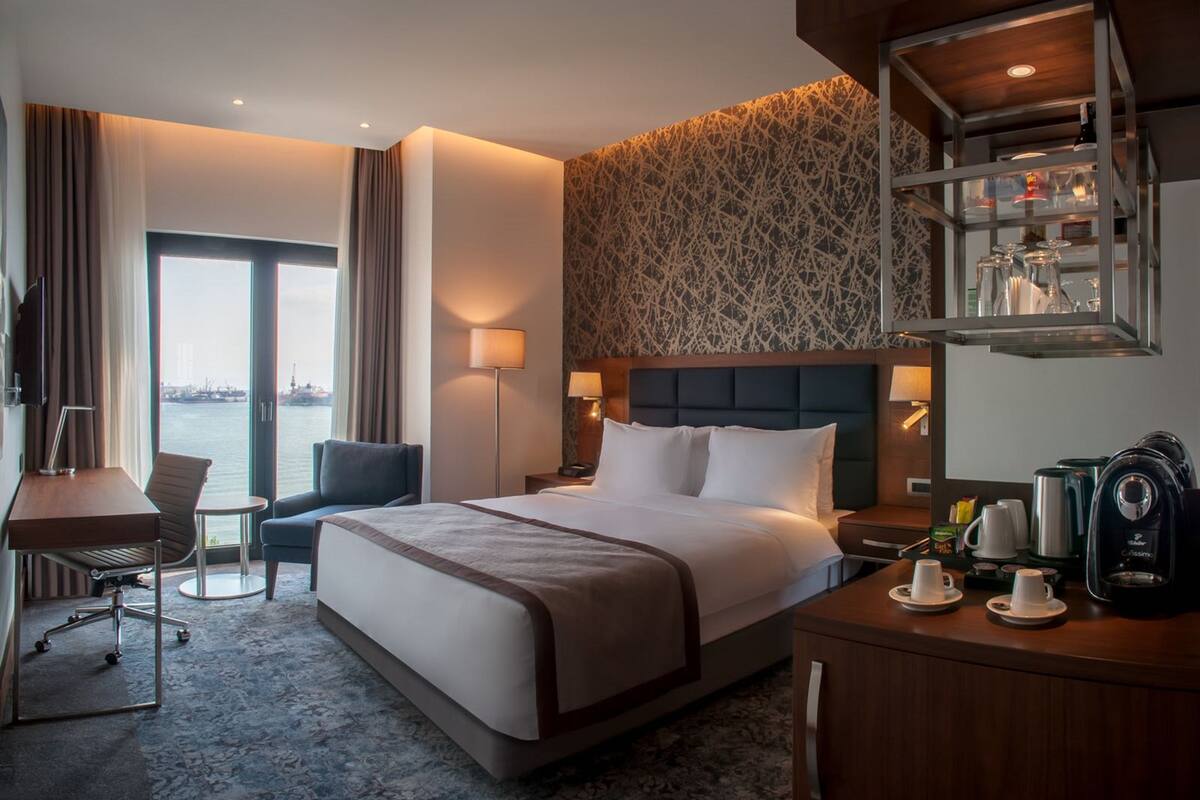 A hotel room with a large bed, a desk, a chair, a small table, and a view of the water.