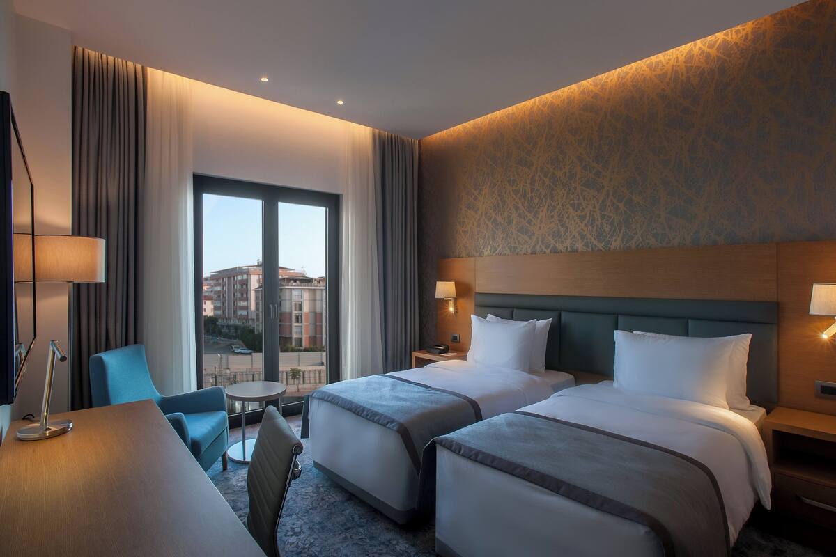A hotel room with two beds, a desk, a chair, a lamp, and a view of buildings.