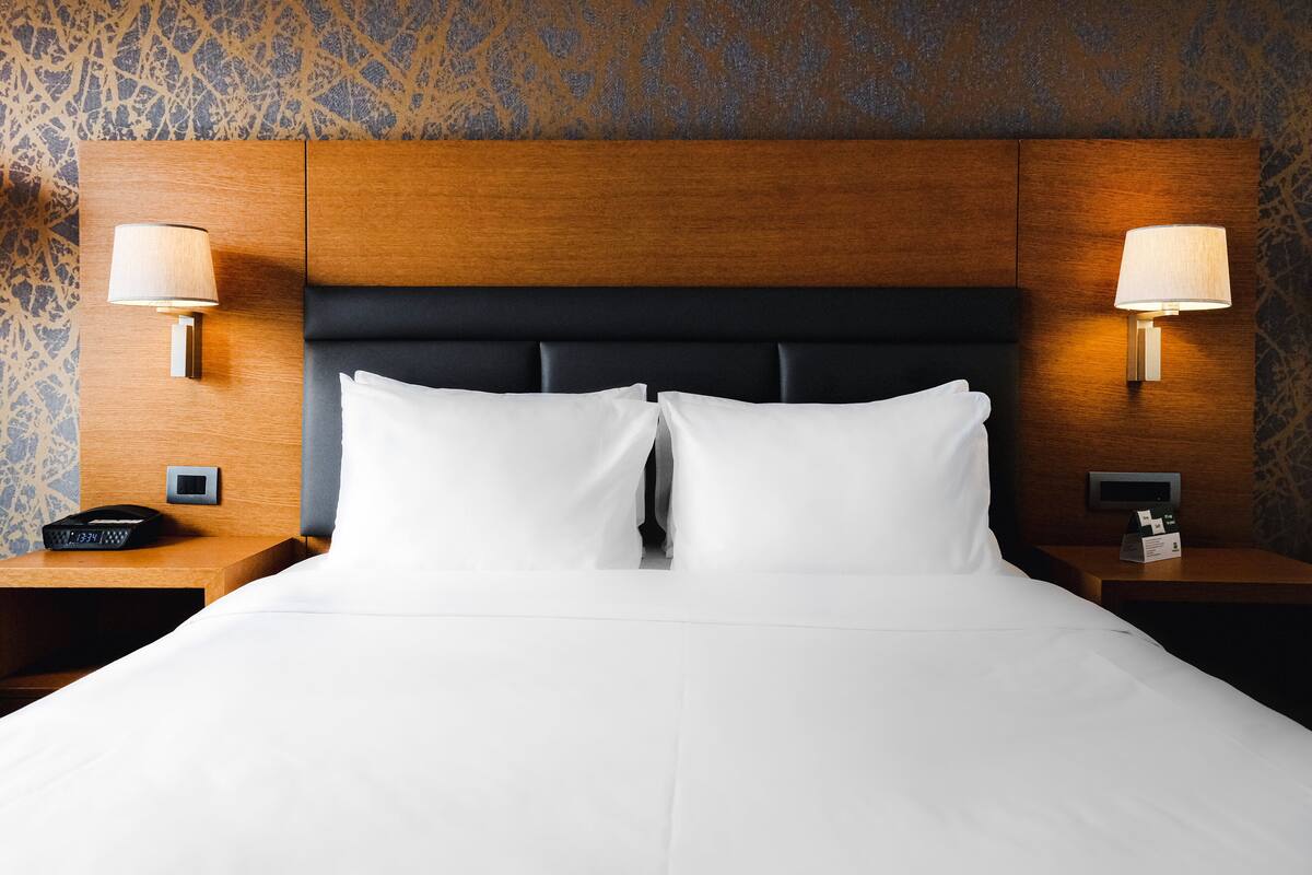 A hotel room with a wooden headboard, two bedside lamps, a digital clock, and a bedside table with a telephone.