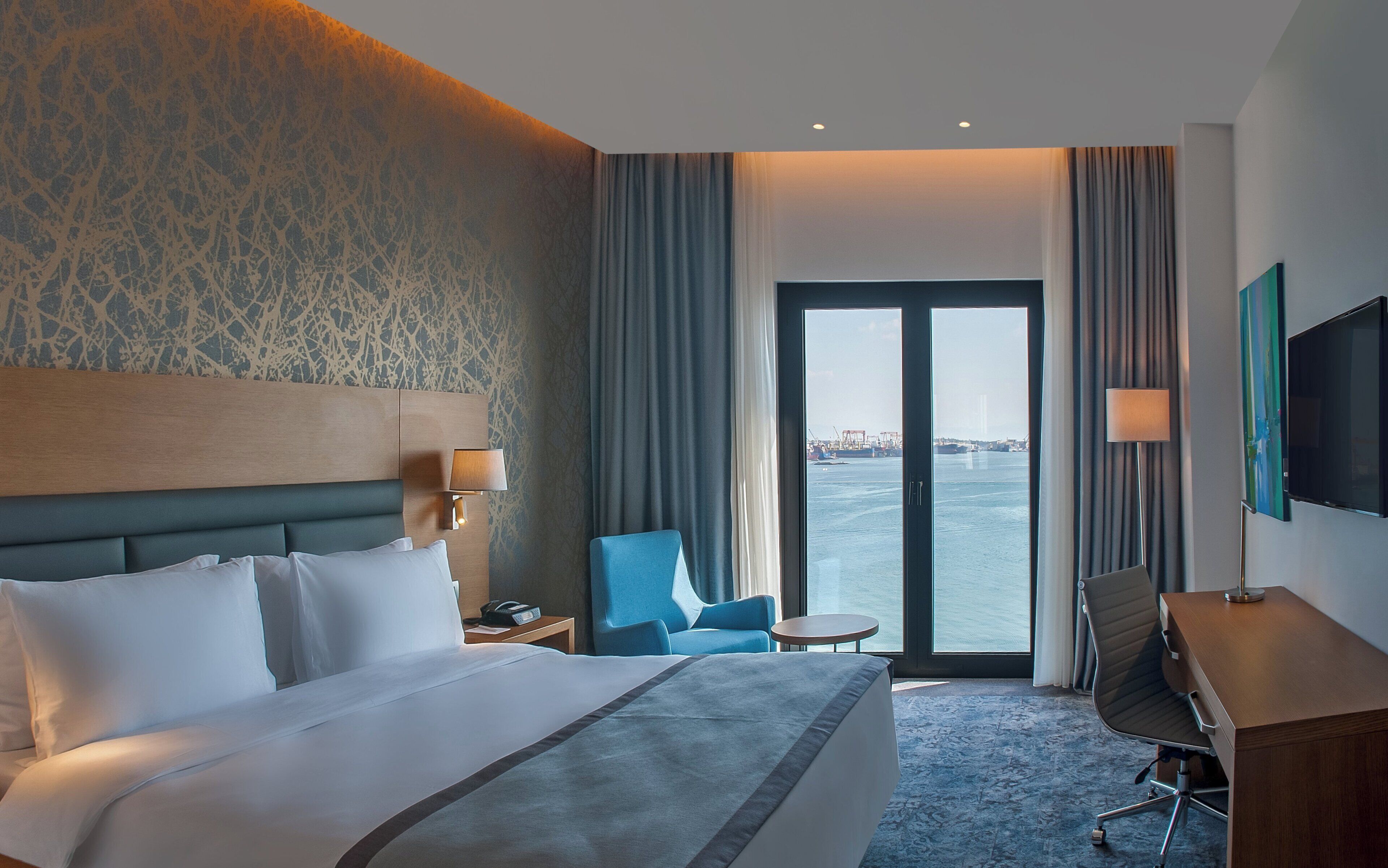 A hotel room with a large bed, a desk, a chair, a TV, and a view of the water.