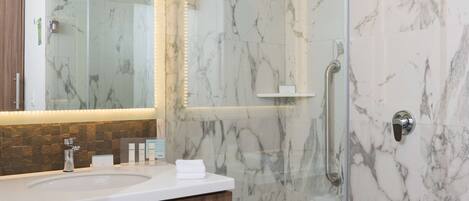 Un baño moderno con ducha de mármol, tocador con puerta de vidrio y repisa de madera con toallas.