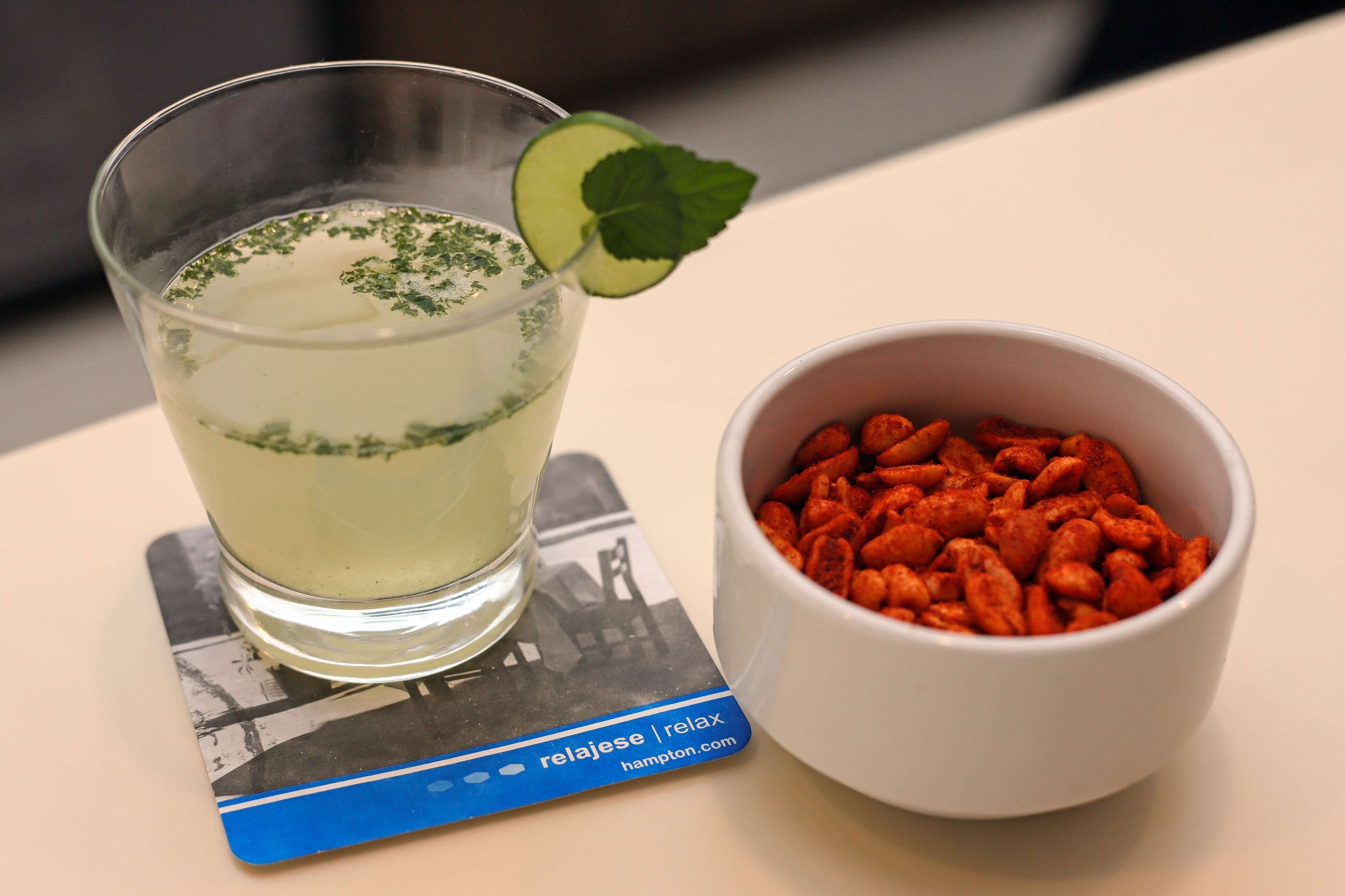 Un vaso de una bebida de color claro con una ramita de menta, un cuenco de bocadillos rojos y un posavasos con una dirección web.