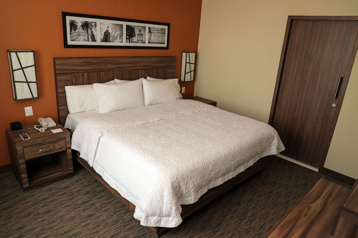 Habitación de hotel con una cama grande, una mesita de noche de madera con un teléfono y un control remoto, y una puerta a la derecha.