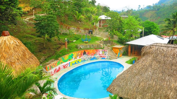 Una piscina con un mural colorido, rodeada de exuberante follaje y una estructura con techo de paja.