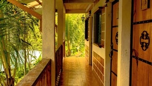 Un pasillo de madera con barandilla de madera, suelo de baldosas y vistas a follaje tropical.