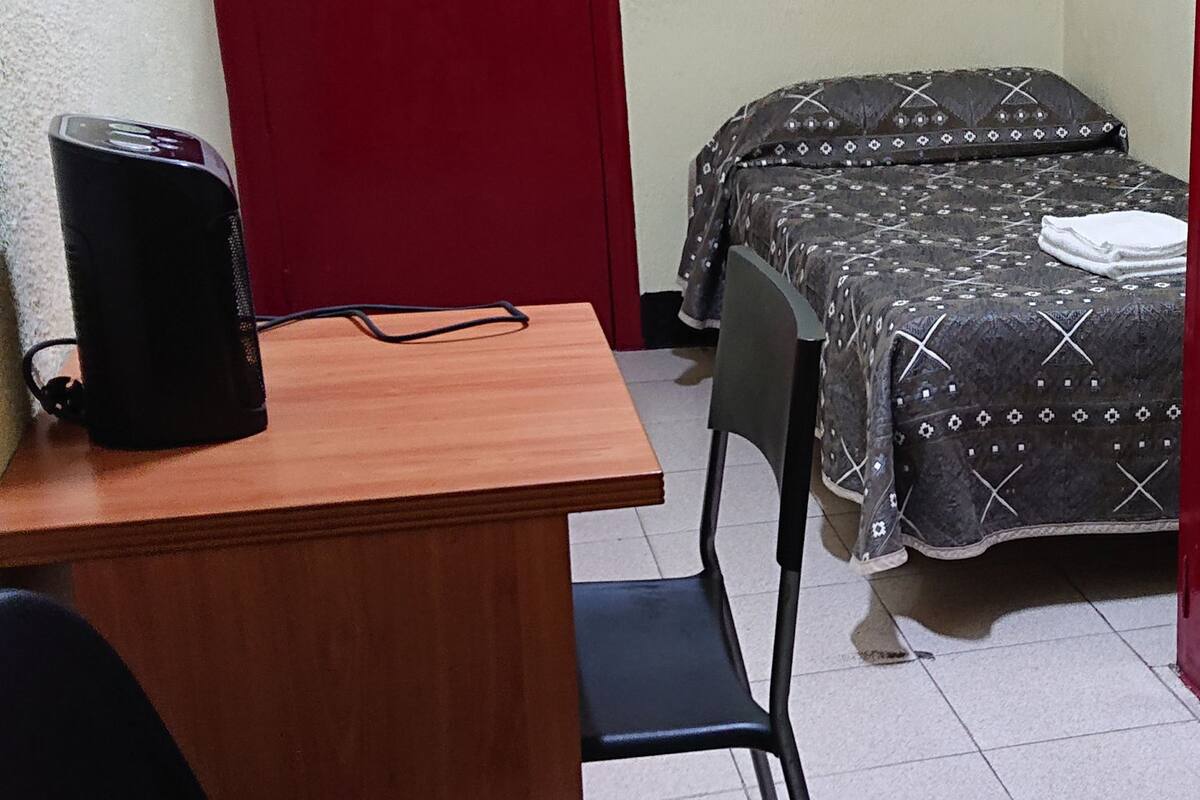 Ein kleines Zimmer mit einem Holztisch, zwei schwarzen Stühlen, einer roten Tür und einem Bett mit gemusterten Textilien.