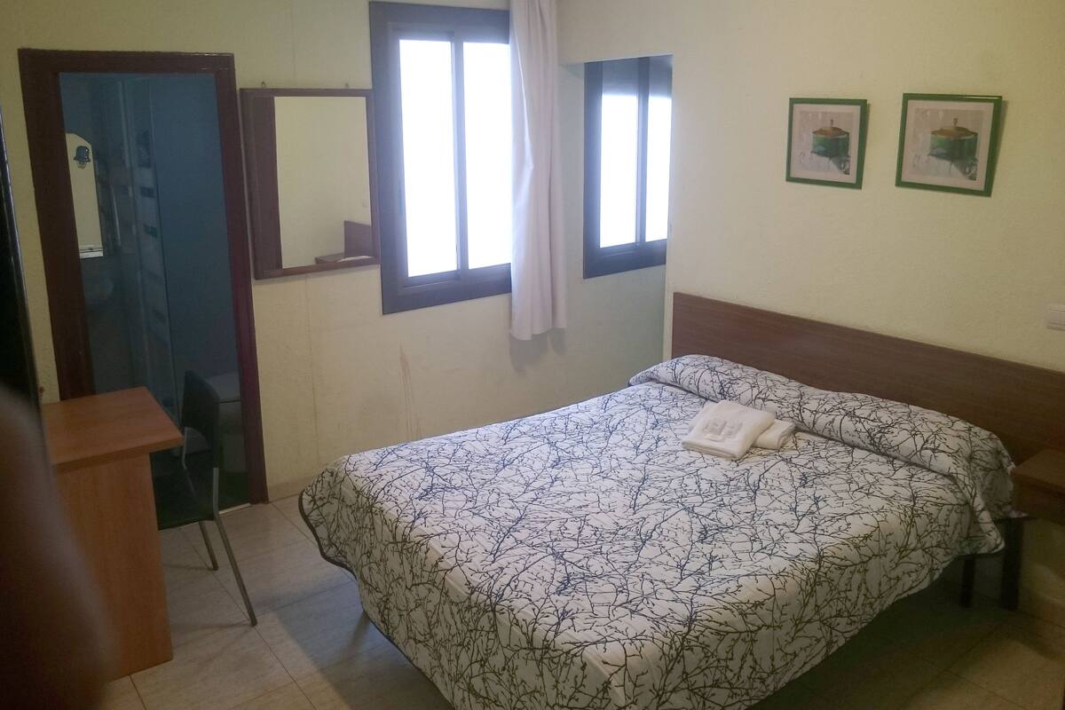 Ein Schlafzimmer mit einem Bett, einem Sessel, einem Schreibtisch, einem Fenster mit Vorhängen und zwei gerahmten Bildern an der Wand.