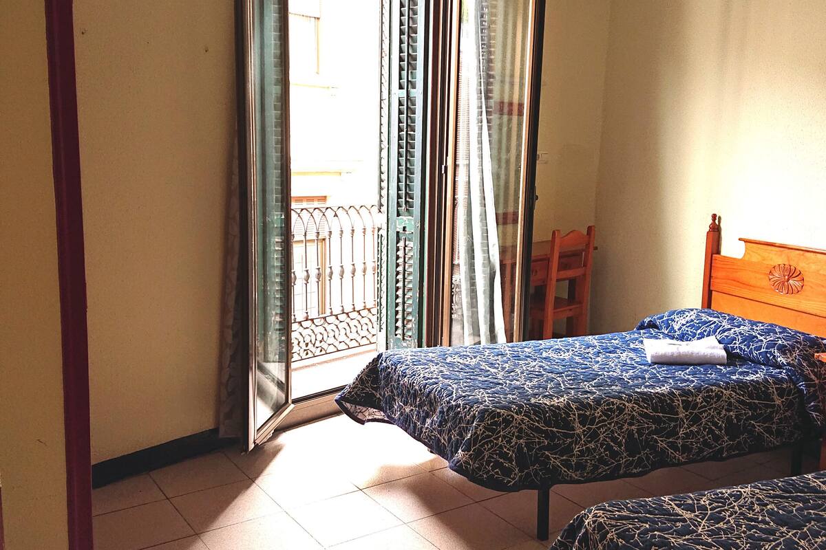 Ein Zimmer mit zwei Betten, einem Holzstuhl und einem Balkon mit Metallgeländer.