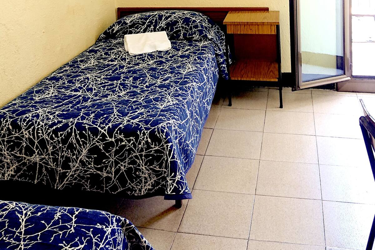 Ein kleines Zimmer mit einem Einzelbett, einer schlichten gelben Wand, einem gerahmten Bild und einer rechtsseitigen Schiebetür.