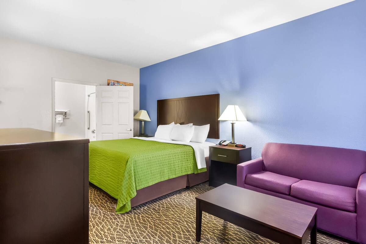 A hotel room with a bed, a purple sofa, a nightstand with a lamp, and a desk.