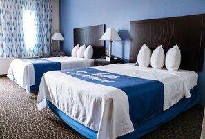 Habitación de hotel con dos camas, cada una con una colcha azul y blanca, almohadas blancas y una mesita de noche con lámpara.