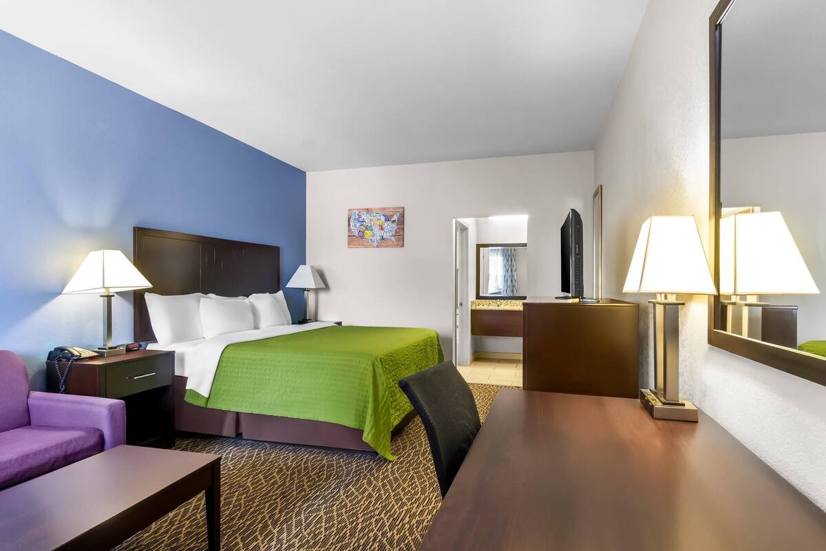 A hotel room with a bed, a desk, a chair, a sofa, a lamp, and a mirror.