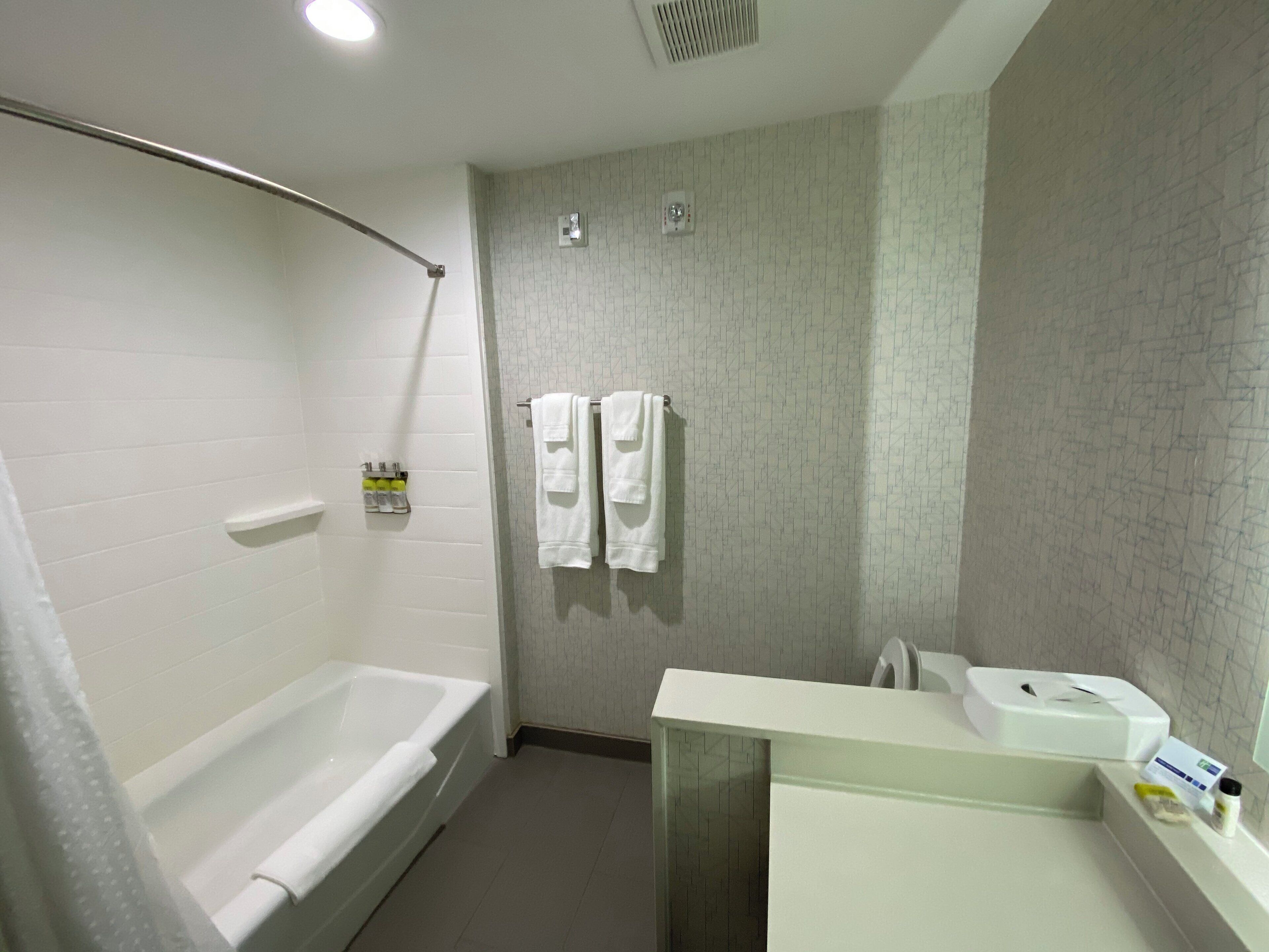 A bathroom with a bathtub, a shower, and a sink with a mirror. There are towels hanging on the wall and a hand sanitizer dispenser.