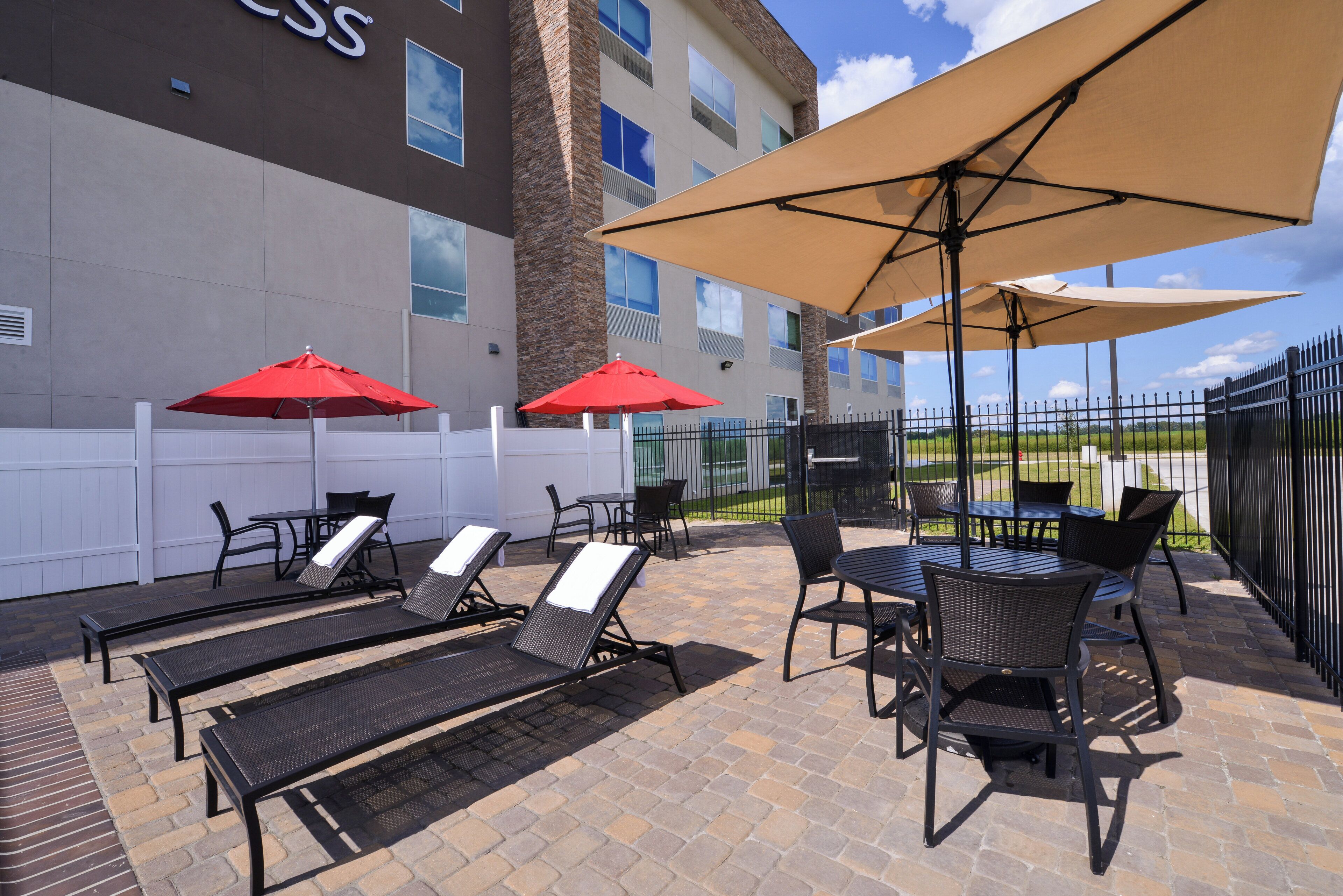 An outdoor patio with lounge chairs and tables, a white fence, and a building with a stone pillar and windows.