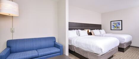 Ett hotellrum med en blå soffa, ett litet bord, två sängar med vita sängkläder, en sänggavel, en väggmonterad lampa och en inramad tavla.