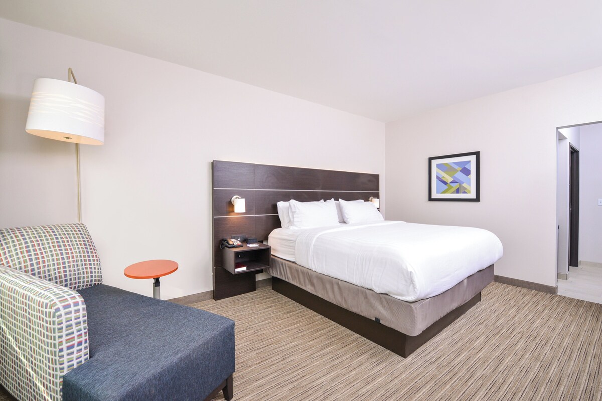A hotel room with a bed, a chair, a small table, a nightstand, a lamp, and a framed picture on the wall.
