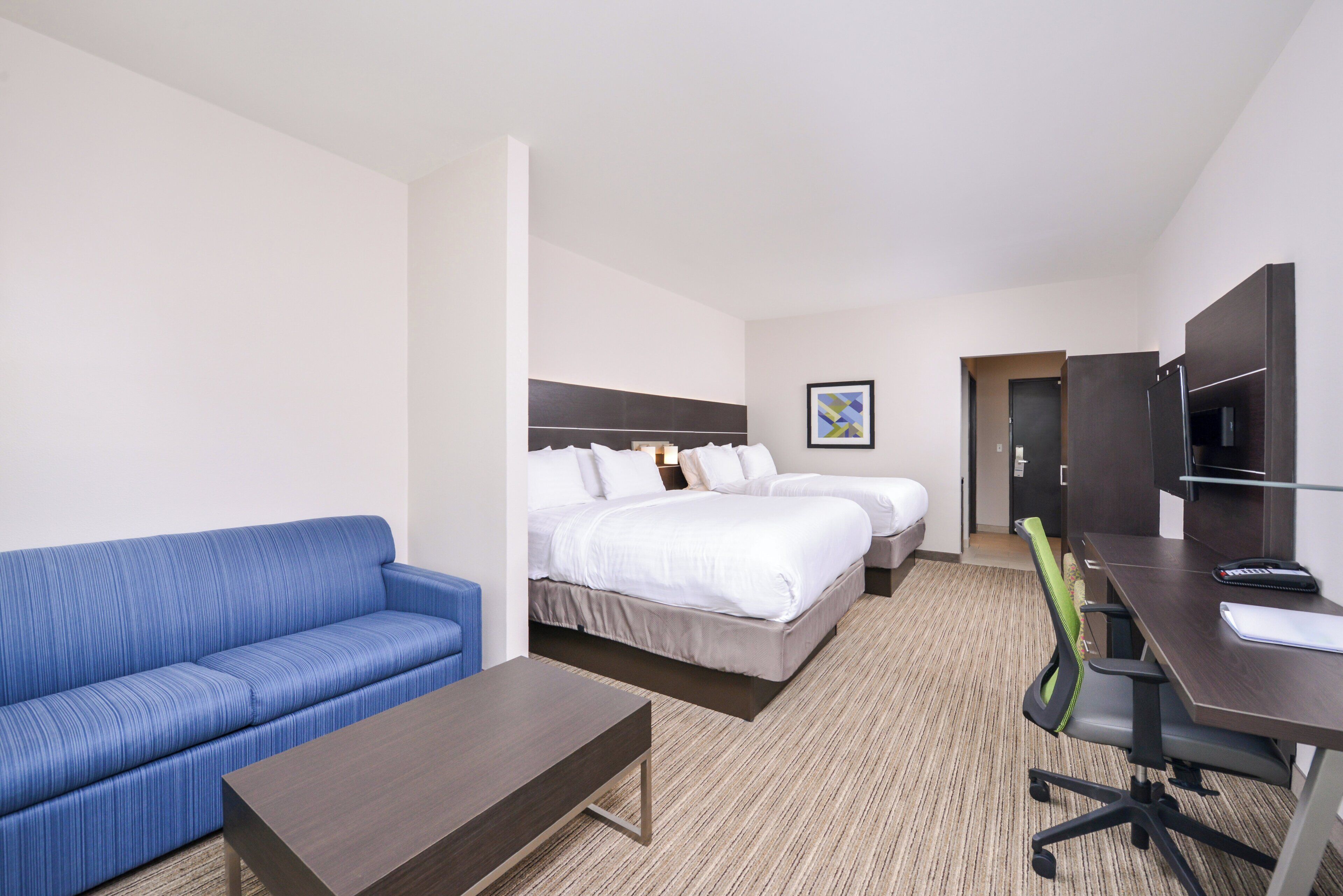 A hotel room with a blue sofa, a single bed, a second bed, a desk with a chair, and a television.