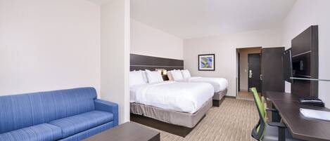 A hotel room with a blue sofa, a single bed, a second bed, a desk with a chair, and a television.